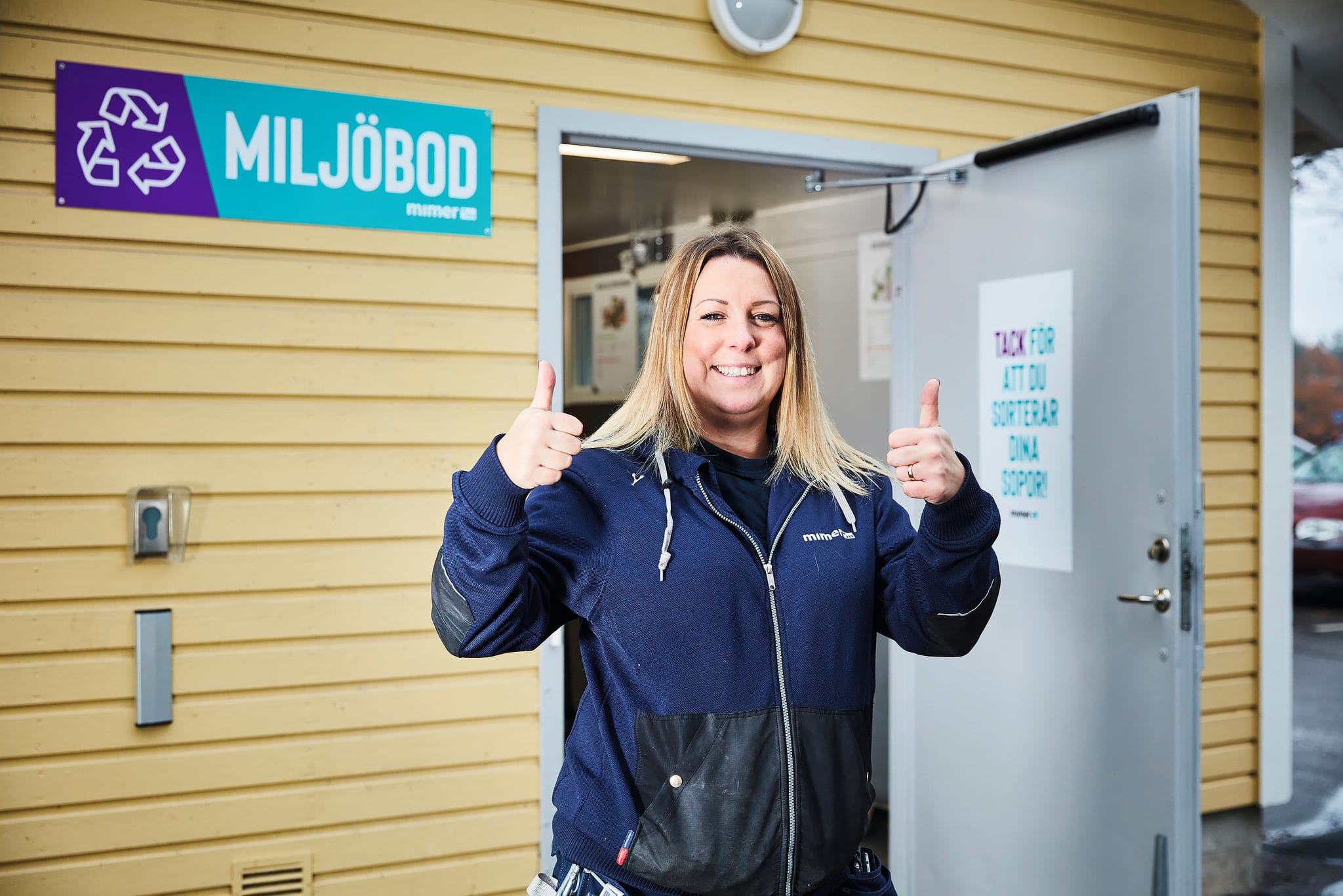 En av våra kvartersvärdar står utanför en miljöbod och gör tummarna upp och har ett strålande leende. I miljöboden kan våra hyresgäster sortera det mesta av sina sopor.